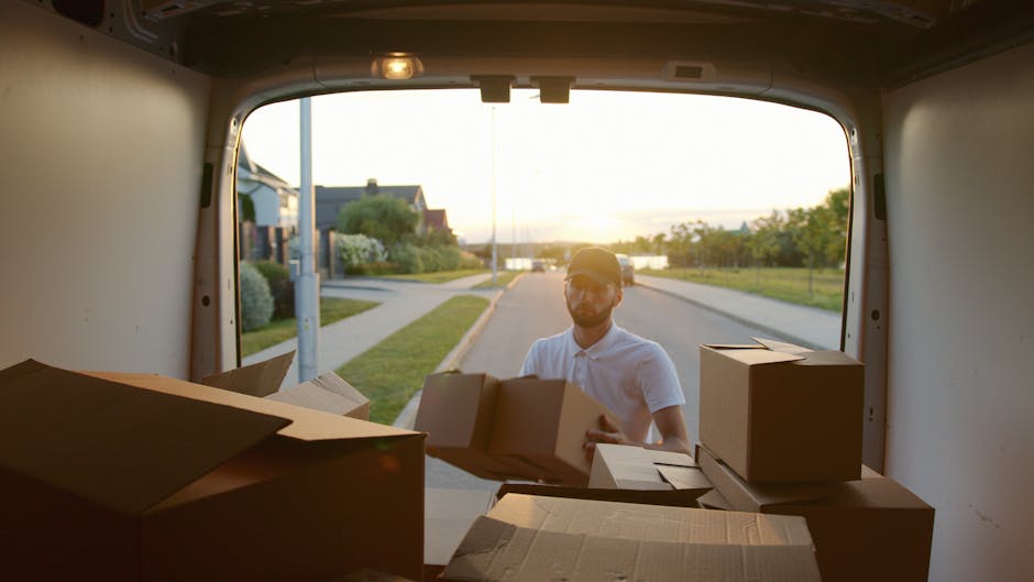 A man wearing a white polo shirt and glasses is standing behind the open rear door of a white moving van during daylight. He is in the process of loading or unloading cardboard boxes of various sizes,
