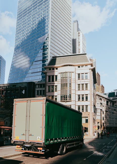 A man wearing a white polo shirt and glasses is standing behind the open rear door of a white moving van during daylight. He is in the process of loading or unloading cardboard boxes of various sizes,