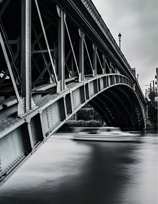 A black and white photograph of a large metal bridge spanning a river, with its detailed steel framework and riveted beams visible beneath the arch. The bridge features decorative lamp posts along its edge, and the water below appears calm with slight ripples. In the background, the sky is overcast, creating a moody atmosphere. This structural element may be part of a cityscape involved in a home relocation or furniture transport process, with the image capturing a scene relevant to moving logistics in the Barnes area. Man With a Van Barnes specializes in removals and moving services, which often include navigating iconic bridges during furniture transport or packing and moving procedures.