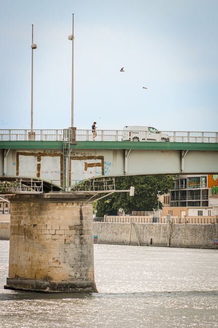 A red delivery van, branded with Coca-Cola, is parked on the upper level of a modern, arched steel bridge spanning a river in Barnes. The bridge features multiple large, curved steel arches with open latticework, painted in a light colour, supporting the roadway. The van is positioned near the centre of the bridge's deck, which is lined with red metal railings. Below, the river reflects parts of the bridge structure, with a riverbank and trees visible in the background. In the foreground, a black metal railing runs alongside a paved walkway, which curves gently along the edge of the river. The scene is captured during the daytime with clear skies, and ambient light highlights the metallic sheen of the bridge. This image relates to house removals and furniture transport by illustrating the logistical aspect of moving items across bridges during a local relocation in Barnes, supported by professional services like Man With a Van Barnes.