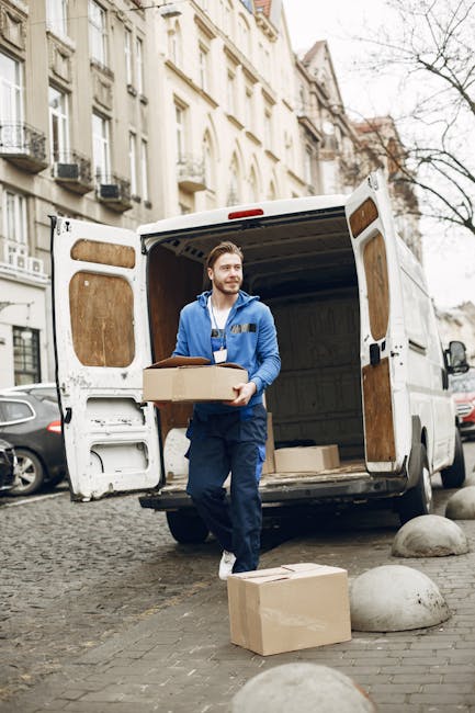A man wearing a blue jacket and dark pants is standing outside a white moving van with its rear doors open on a cobblestone street lined with multi-storey residential buildings. He is carrying a cardboard box, with one matching box placed on the ground nearby. The van's interior shows several other boxes and packing materials, indicating an active home relocation or furniture transport process. The scene is set during daytime, with natural light illuminating the street, and large rounded stone bollards are visible along the pavement. The image depicts the loading process typical of professional removals, illustrating careful handling of packed items in a narrow street environment, relevant to house removals and packing services provided by Man With a Van Barnes.